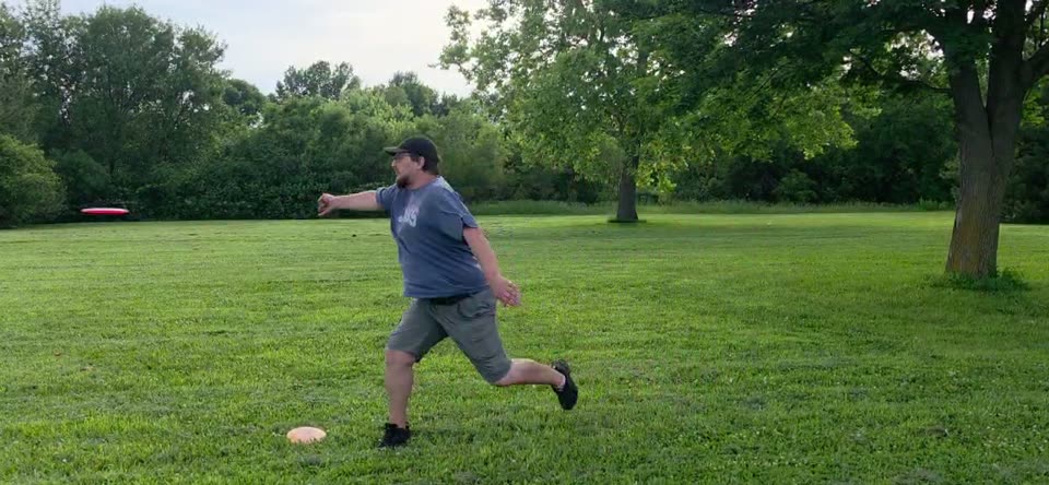 A disc golfer filmed side-on in an open field, front foot planted, disc just released and flying left across the frame